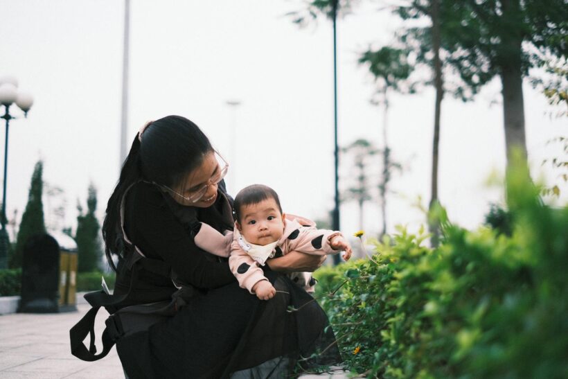 A mother holding her baby in an urban park setting, surrounded by greenery.